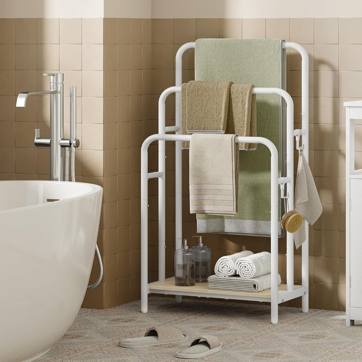 A freestanding, white metal towel rack with three tiers, placed in a bathroom setting next to a bathtub and a wall-mounted towel holder.